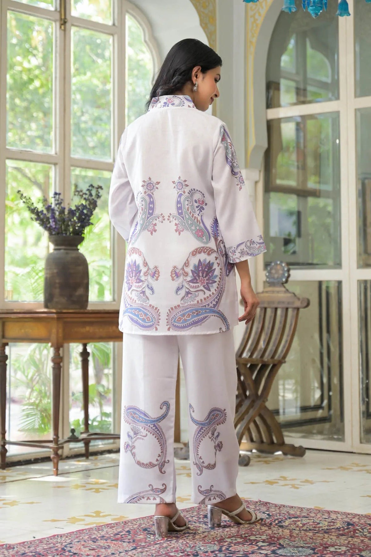 Woman wearing a white outfit with purple and blue patterns in a room with large windows.