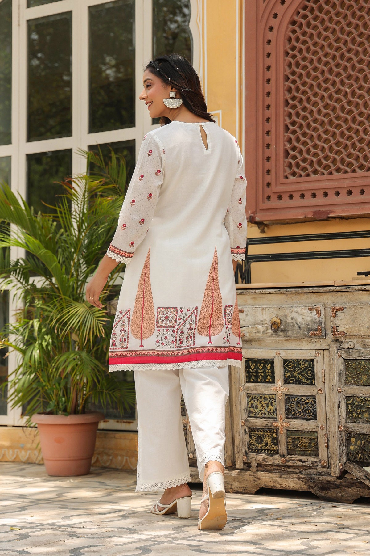 Woman in a white embroidered kurta with red patterns standing in front of a decorative wall.