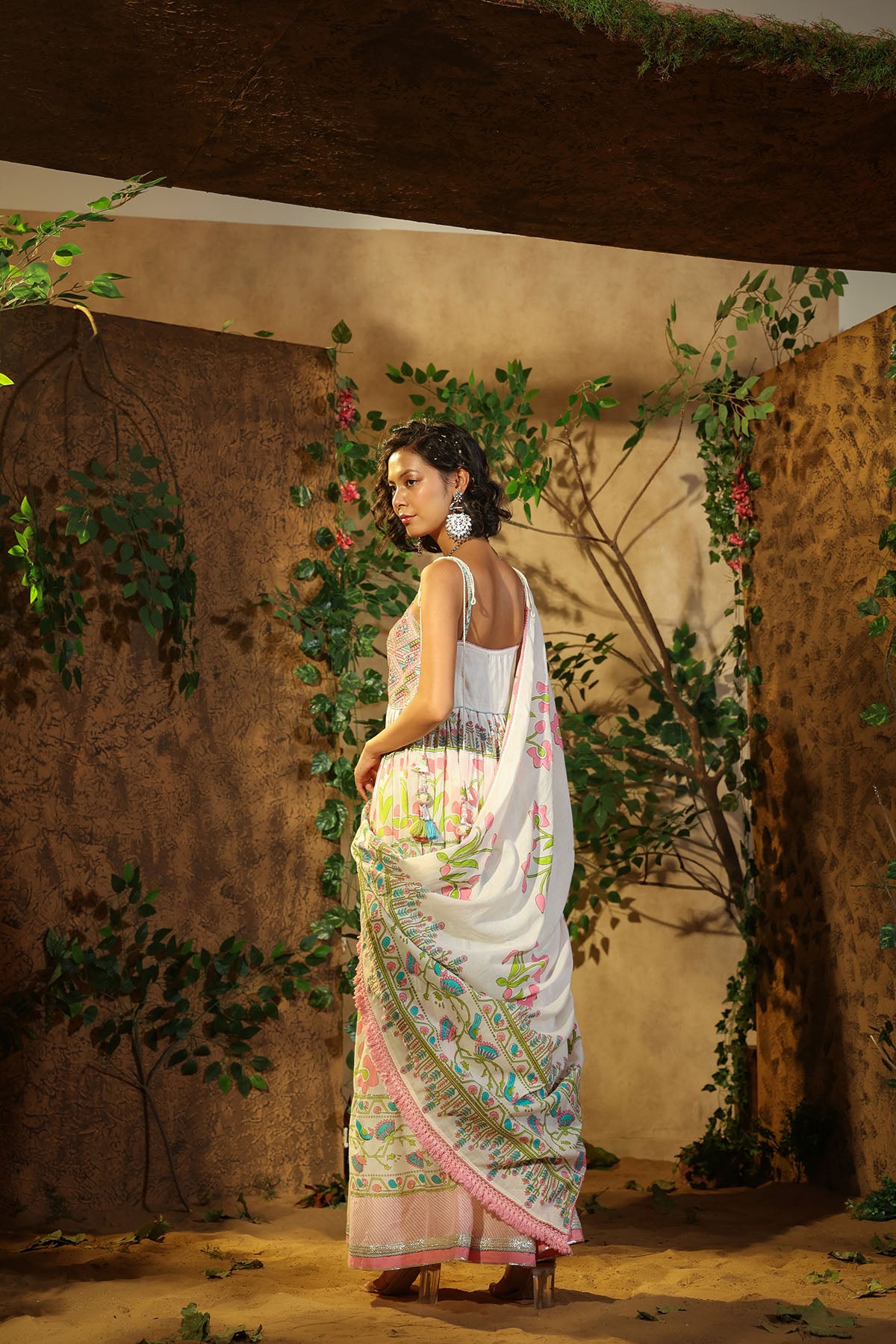 Woman in a floral saree standing in a rustic setting with plants and a textured wall.