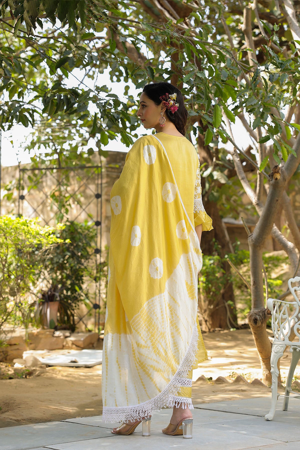 Woman in a yellow saree with white patterns standing outdoors near trees and a bench.