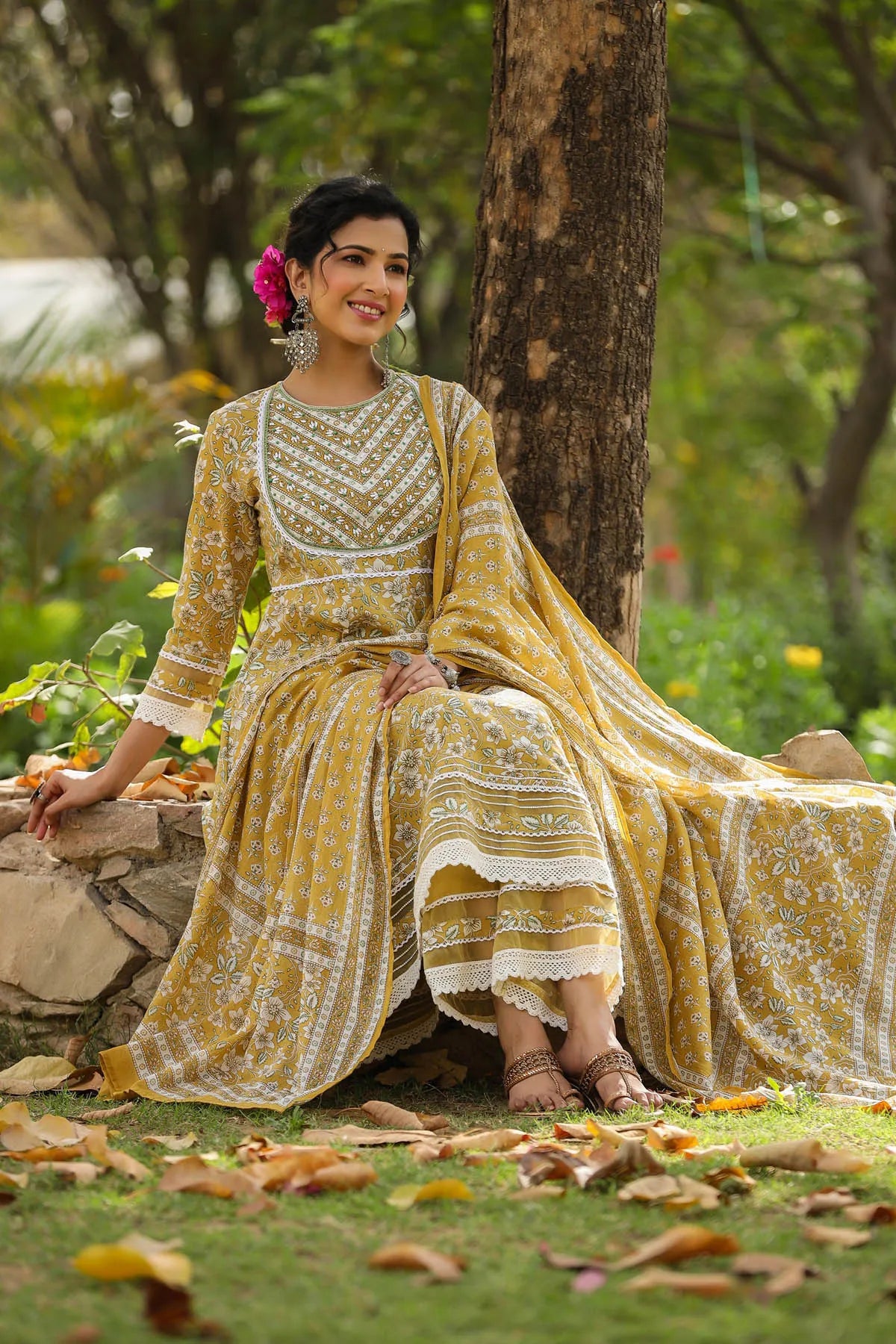 Woman in a yellow and white traditional outfit sitting under a tree with greenery around.