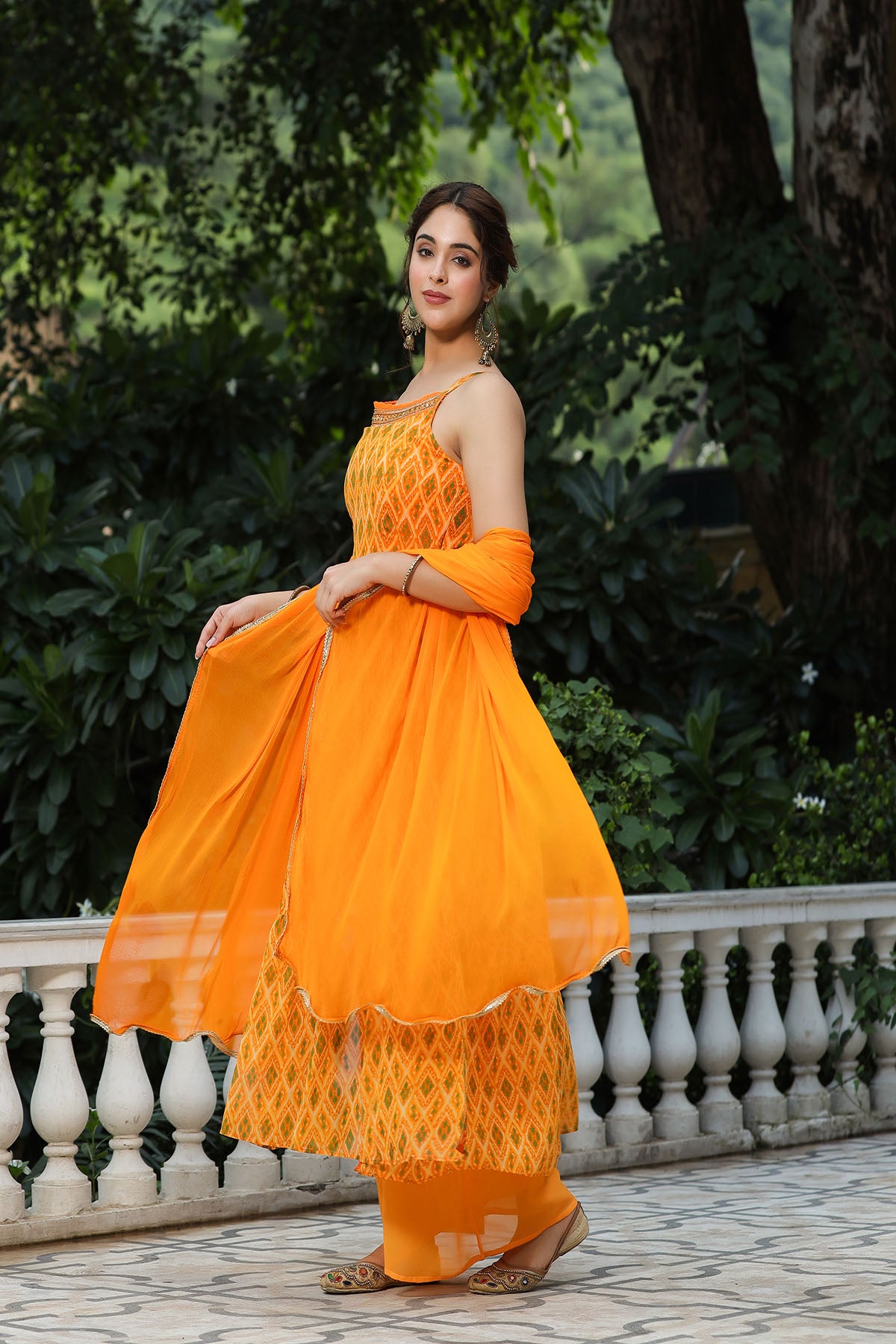 Woman in an orange traditional outfit standing outdoors with greenery in the background