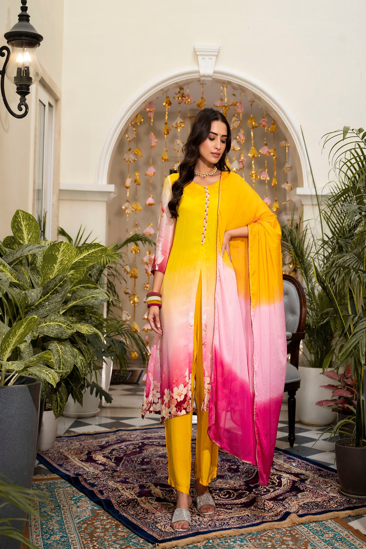 Woman in a yellow and pink outfit standing in an outdoor setting with plants and a decorative arch.