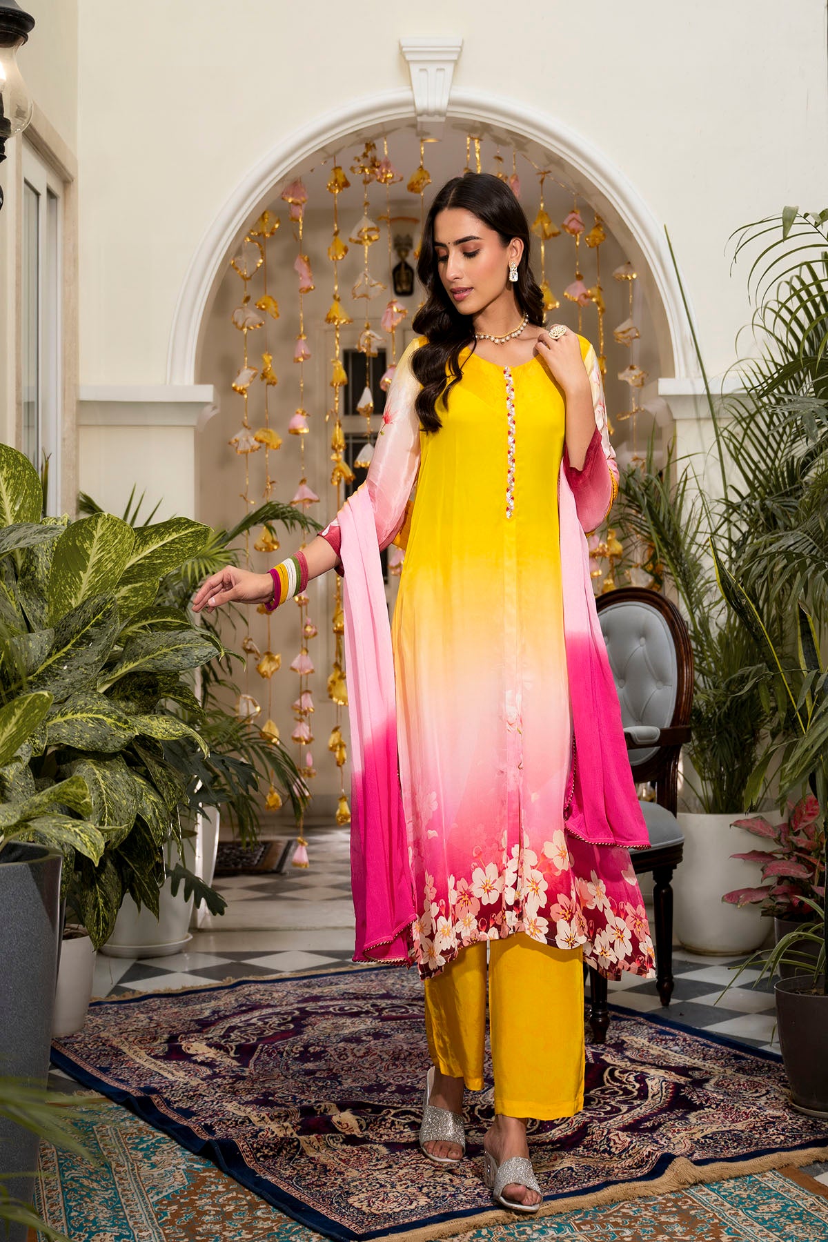 Woman in a yellow and pink traditional outfit standing in an ornate room with plants.