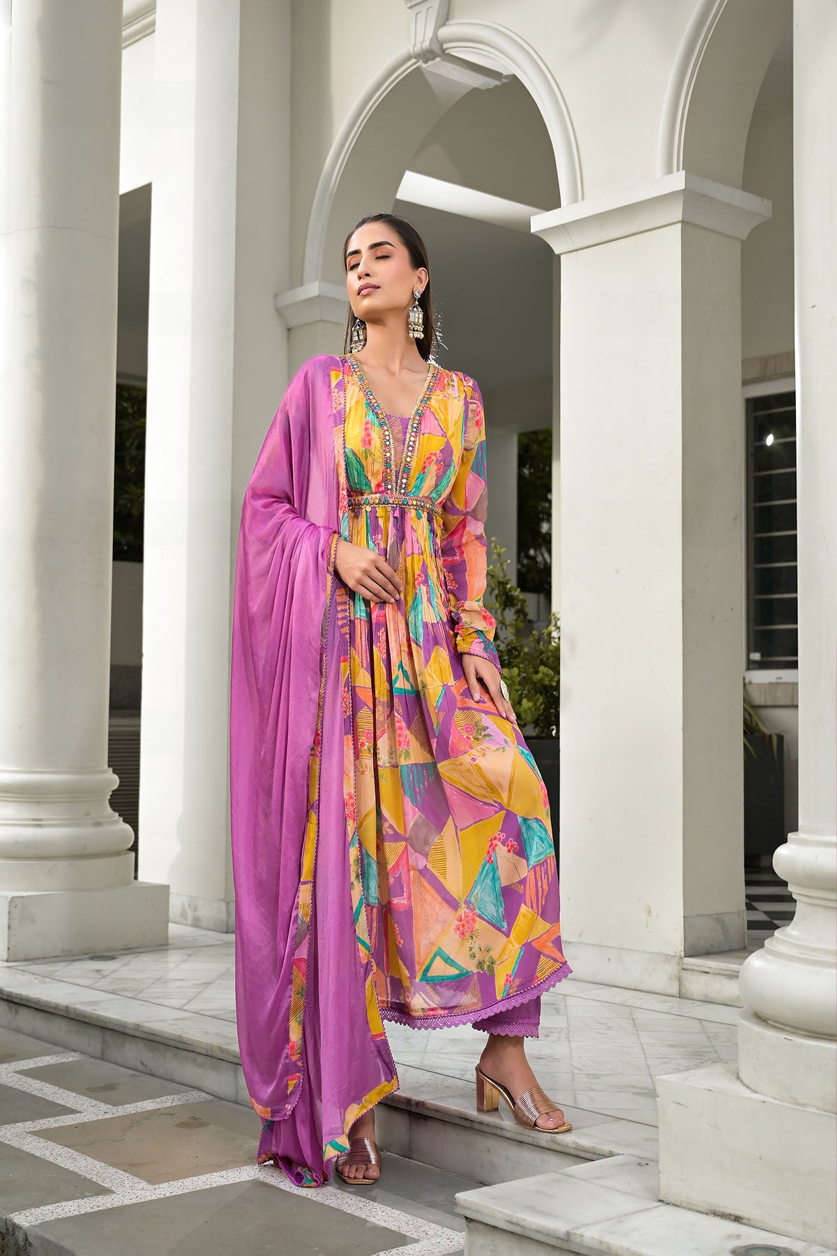 Woman in a colorful dress with a pink dupatta standing in front of classical architecture.