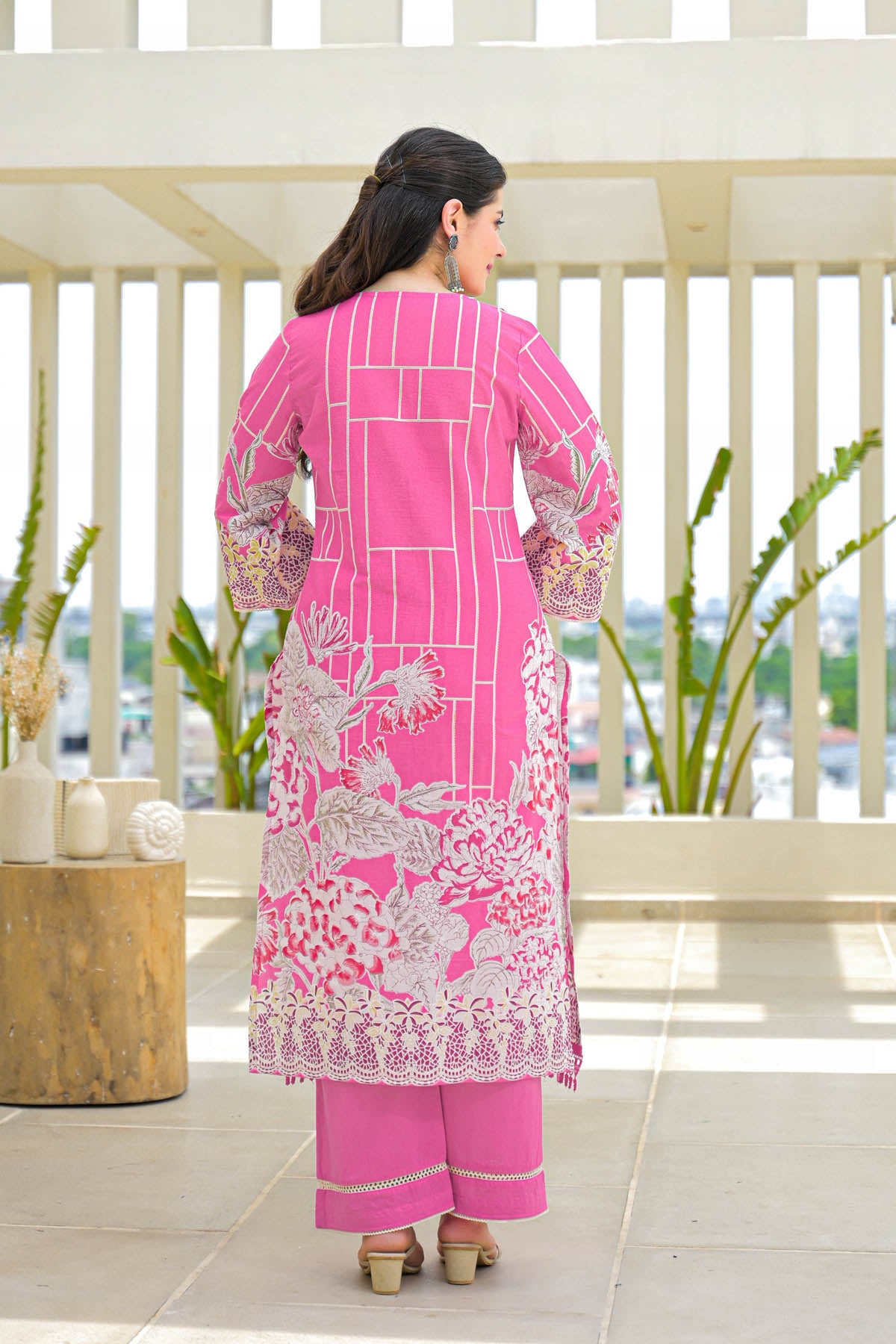 Woman wearing a pink embroidered outfit on a balcony with plants and a table.