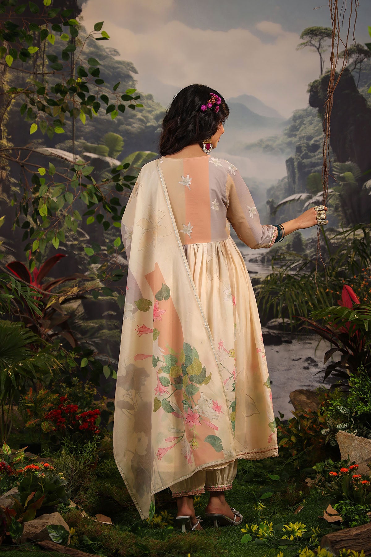 Woman in a floral dress standing in a painted landscape with mountains and trees.
