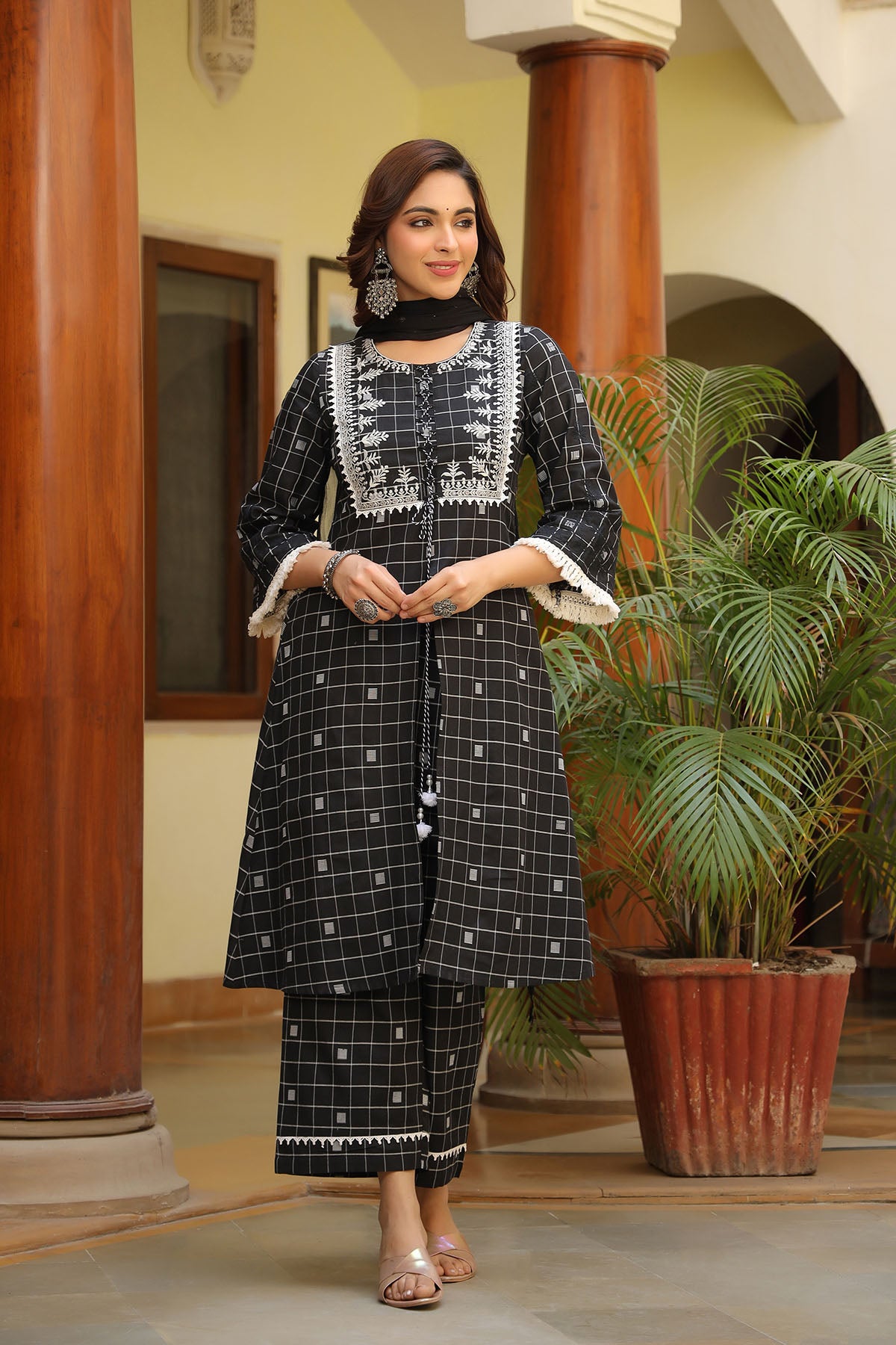 Woman in traditional black and white outfit standing indoors with decorative columns and plants.