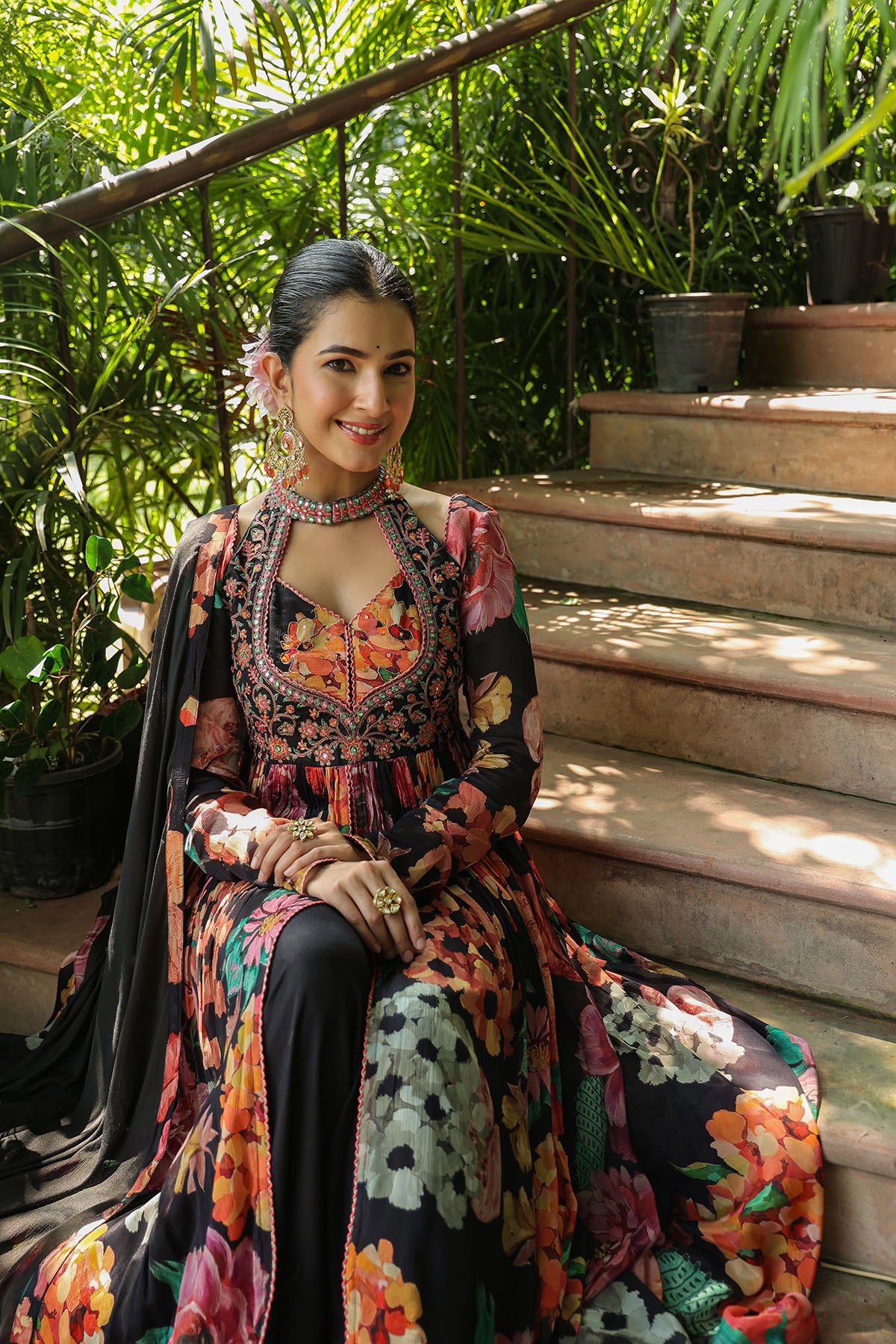 Woman in a colorful traditional outfit sitting on steps with greenery in the background