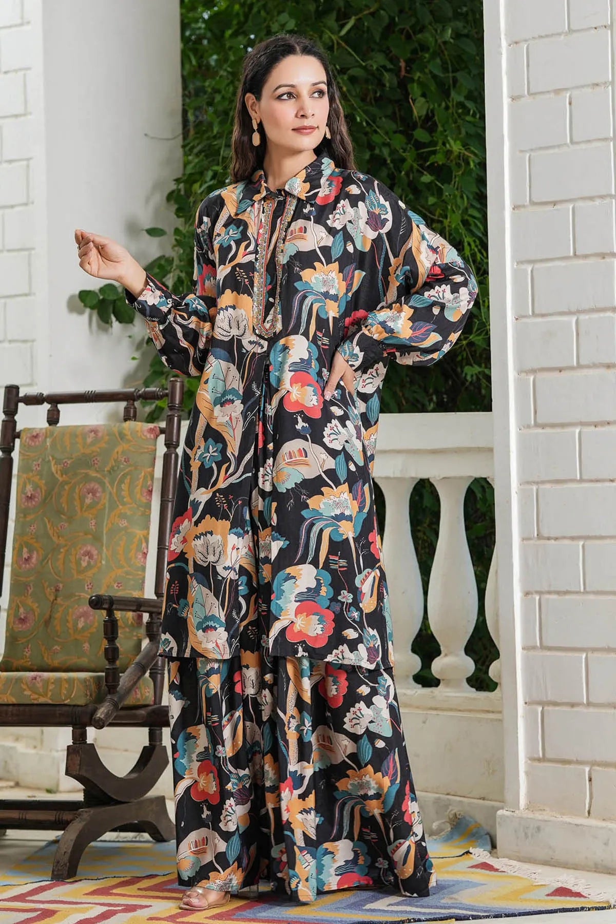 Woman wearing a colorful floral dress standing in a decorated room with a chair and plants.