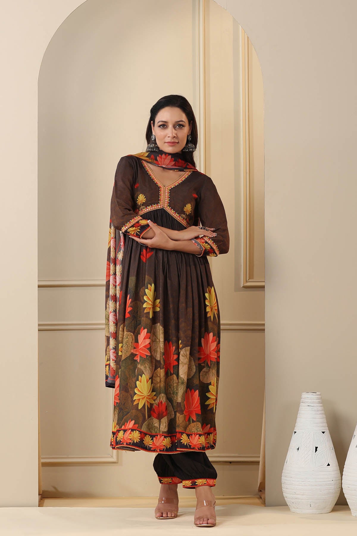 Woman wearing a floral dress standing in a room with a vase and mirror.