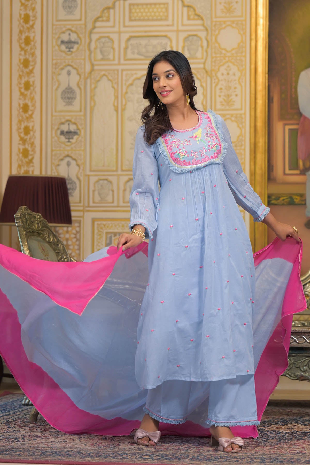 Woman in a light blue traditional outfit with pink dupatta in an ornate room.