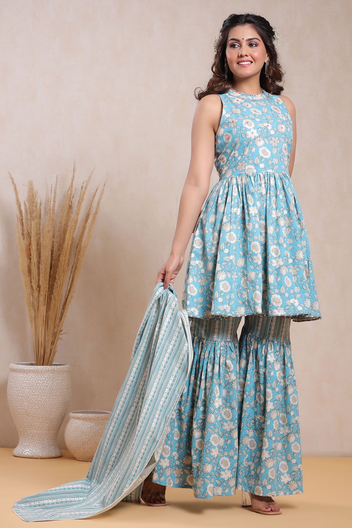 Woman wearing a blue floral dress holding a matching dupatta against a beige background
