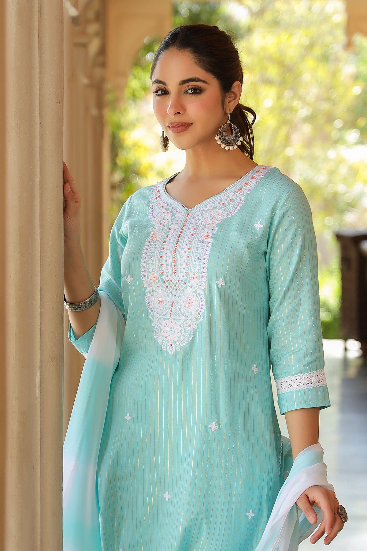 Woman wearing a light blue traditional outfit with white embroidery, standing outdoors.