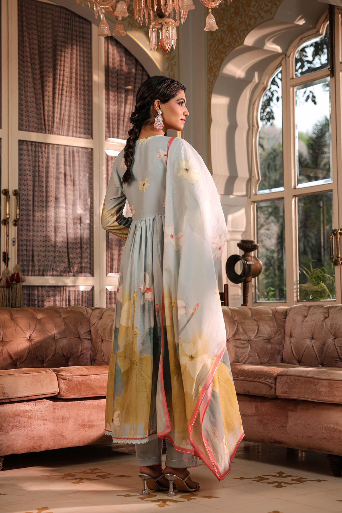 Woman in a light blue and yellow traditional outfit standing in a decorated room with large windows.