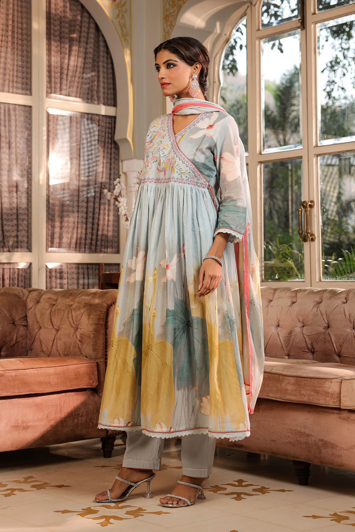Woman in a light blue and yellow traditional outfit standing in a room with large windows and brown furniture.
