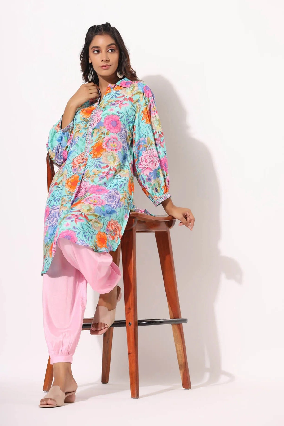 Woman sitting on a stool wearing a colorful floral top and pink pants against a white background