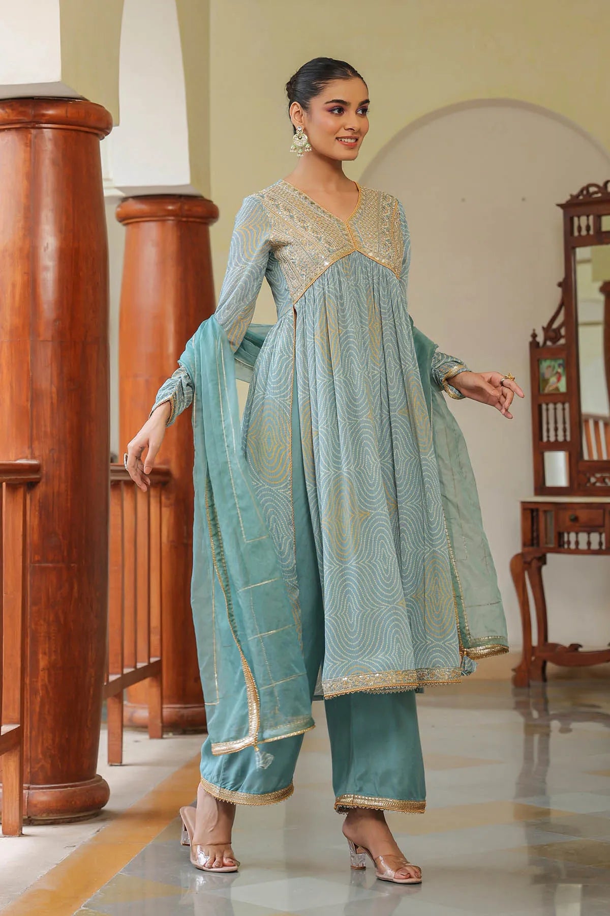 Woman in a light blue traditional outfit standing in a room with wooden columns and a mirror.