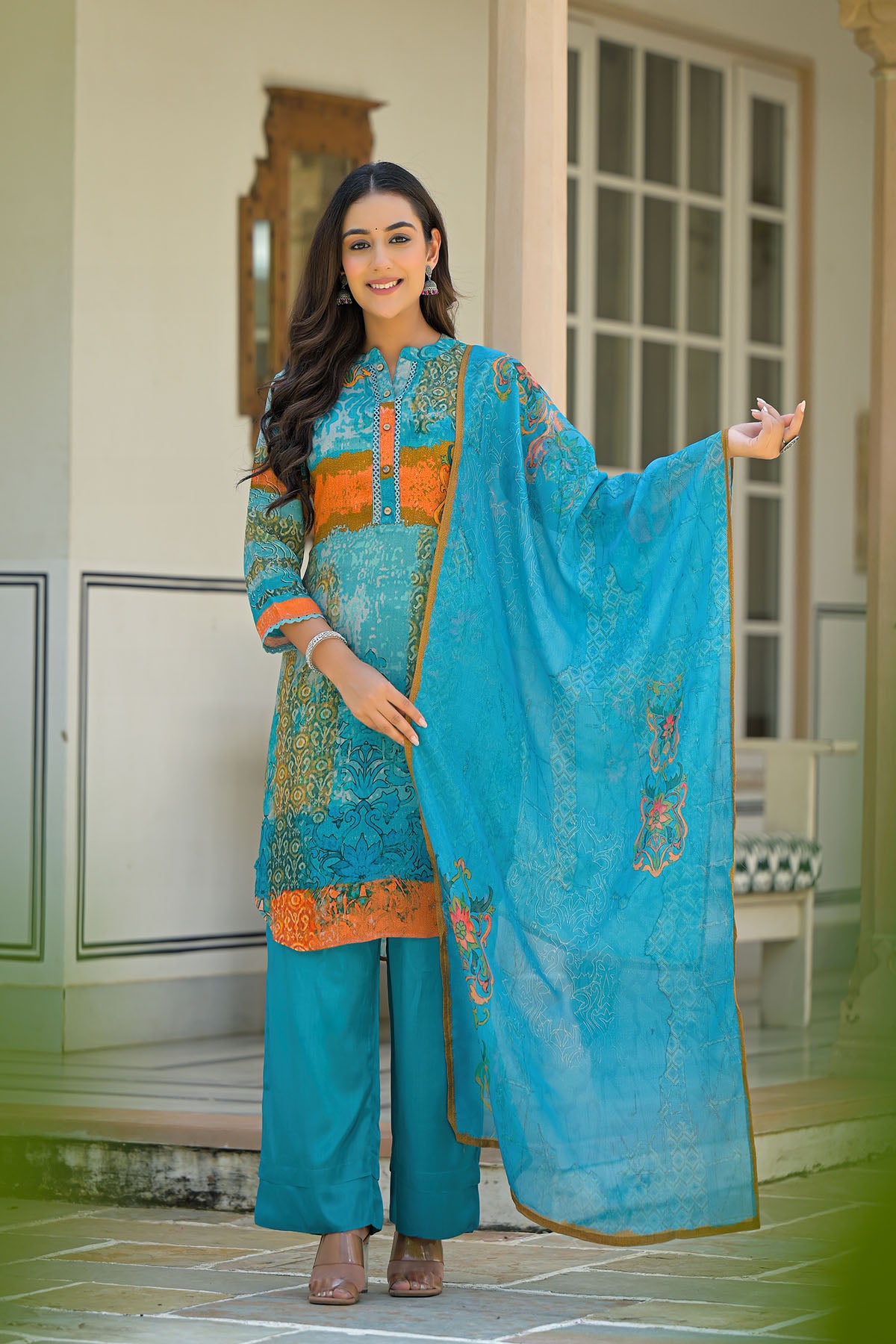 Woman in a turquoise traditional outfit with orange accents, standing indoors.