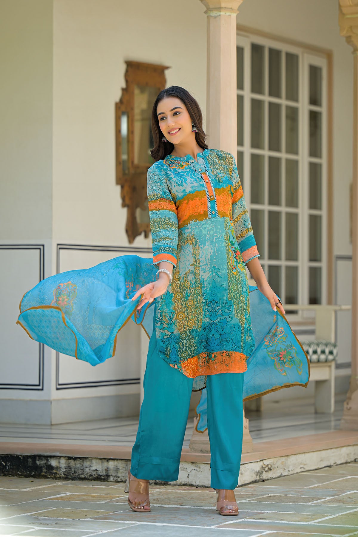 Woman in a turquoise and orange traditional outfit standing in a decorative indoor setting.