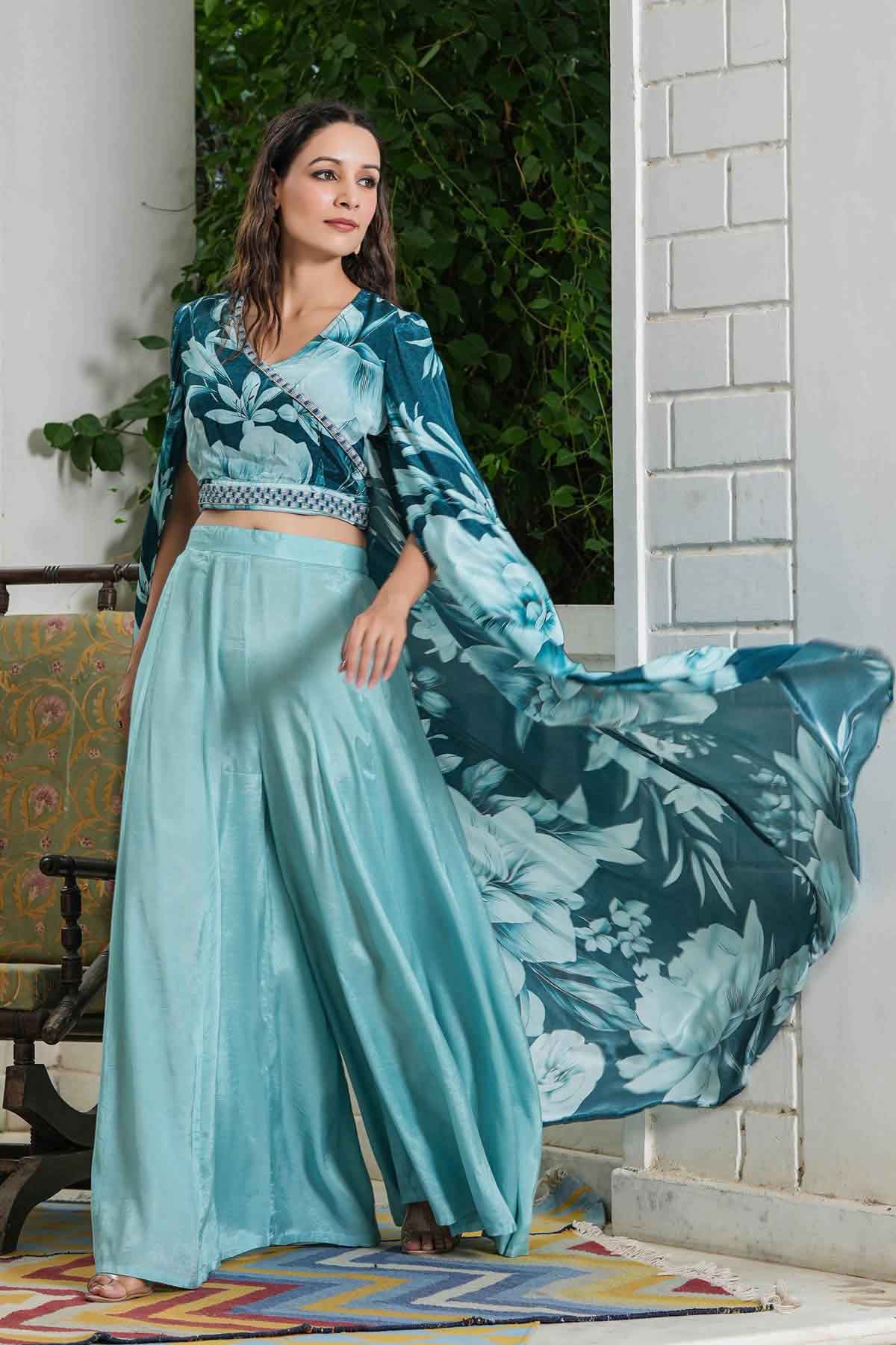 Woman in a teal and blue traditional outfit with a floral shawl, standing indoors.