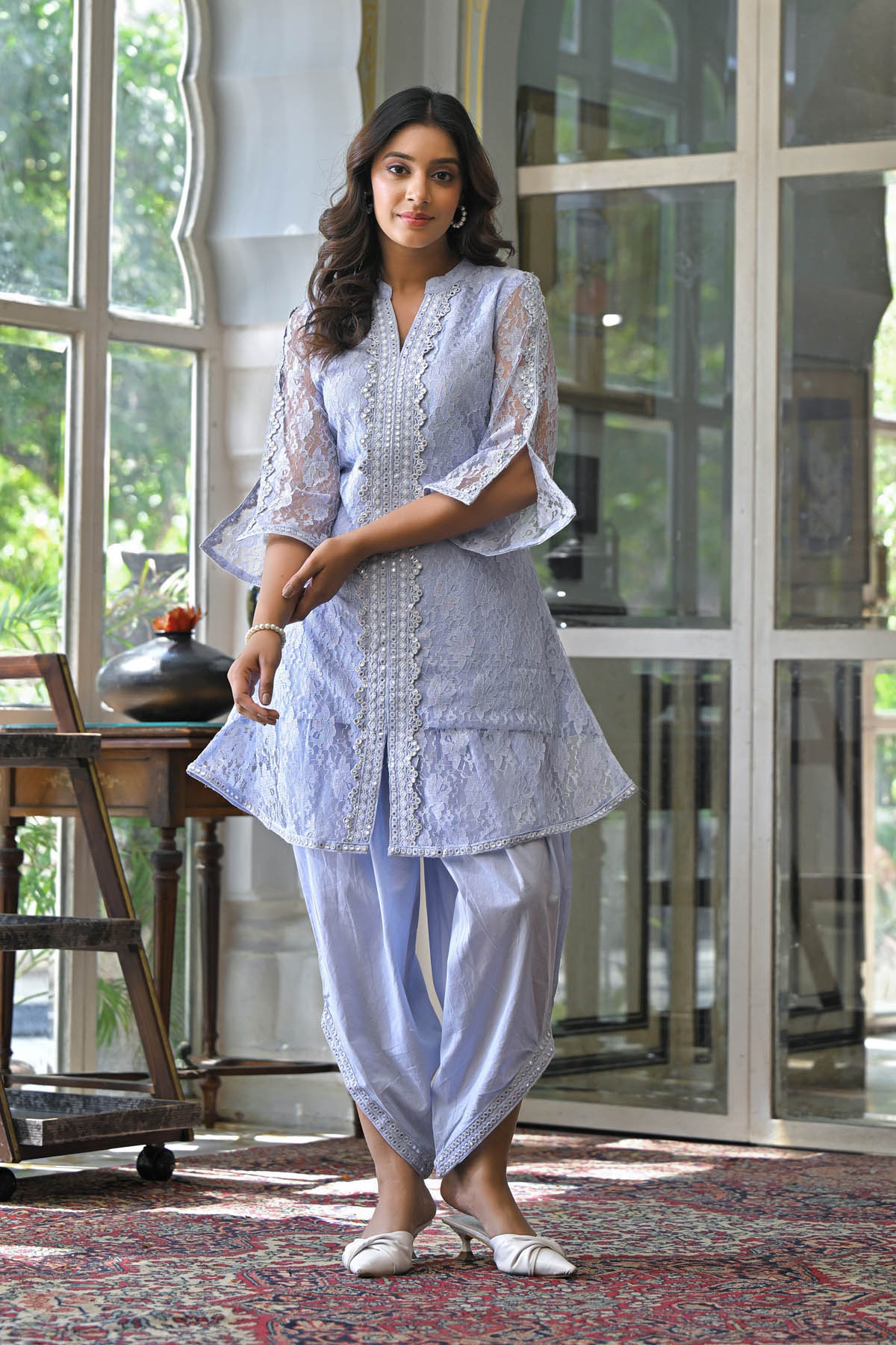 Woman in a light blue traditional outfit standing in a room with large windows.