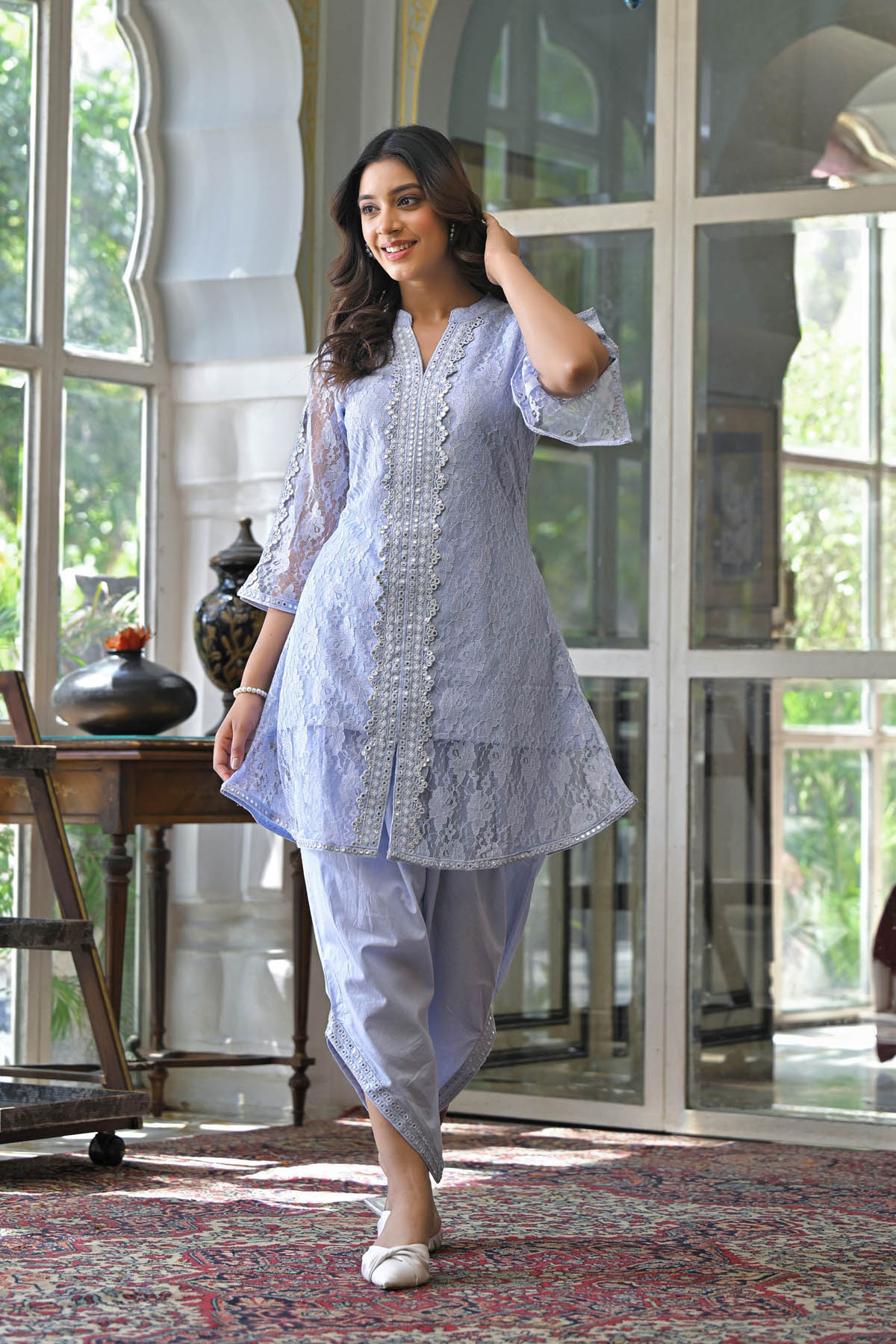 Woman in a light blue traditional outfit standing in a well-lit room with large windows.