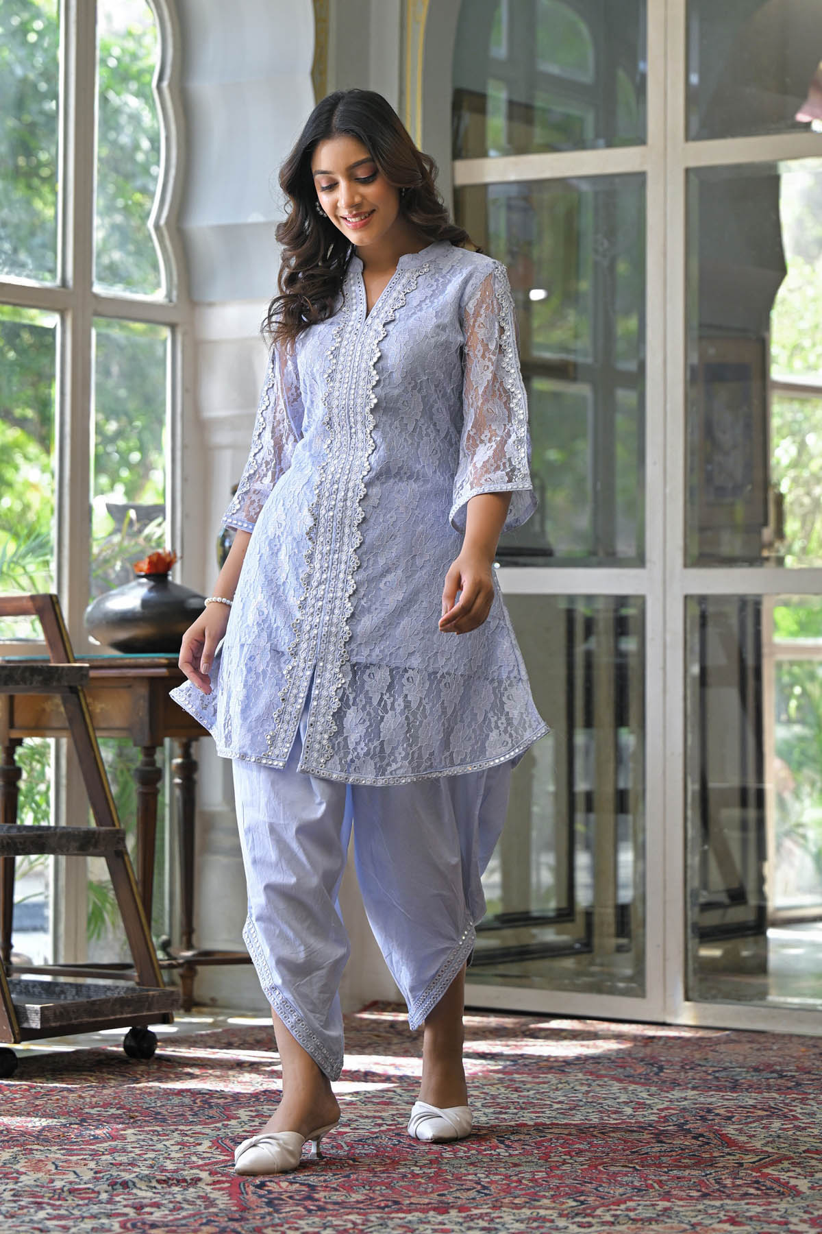 Woman in a light blue traditional outfit walking indoors with large windows in the background