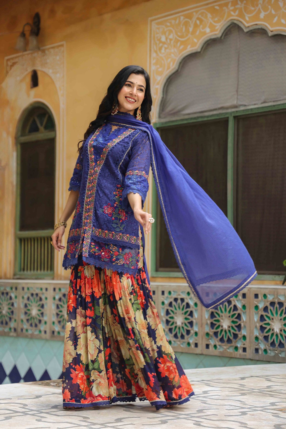 Woman in traditional outfit with a blue dupatta standing in front of an ornate building.