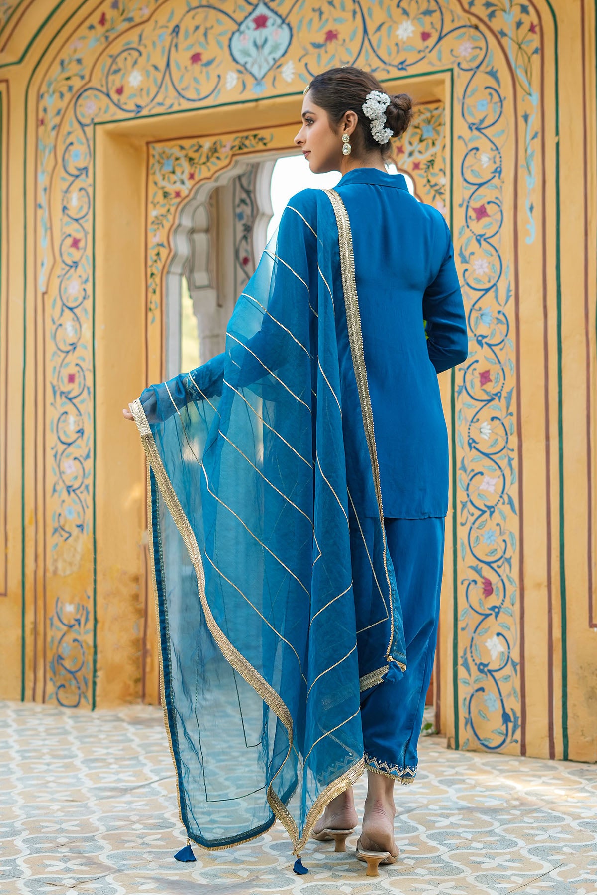 Woman in a blue traditional outfit standing in front of an ornate mirror.