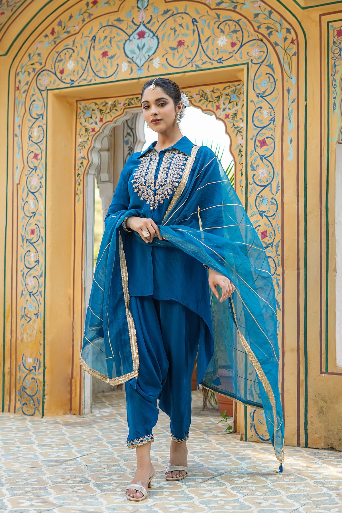 Woman in a blue traditional outfit standing in front of an ornate archway.