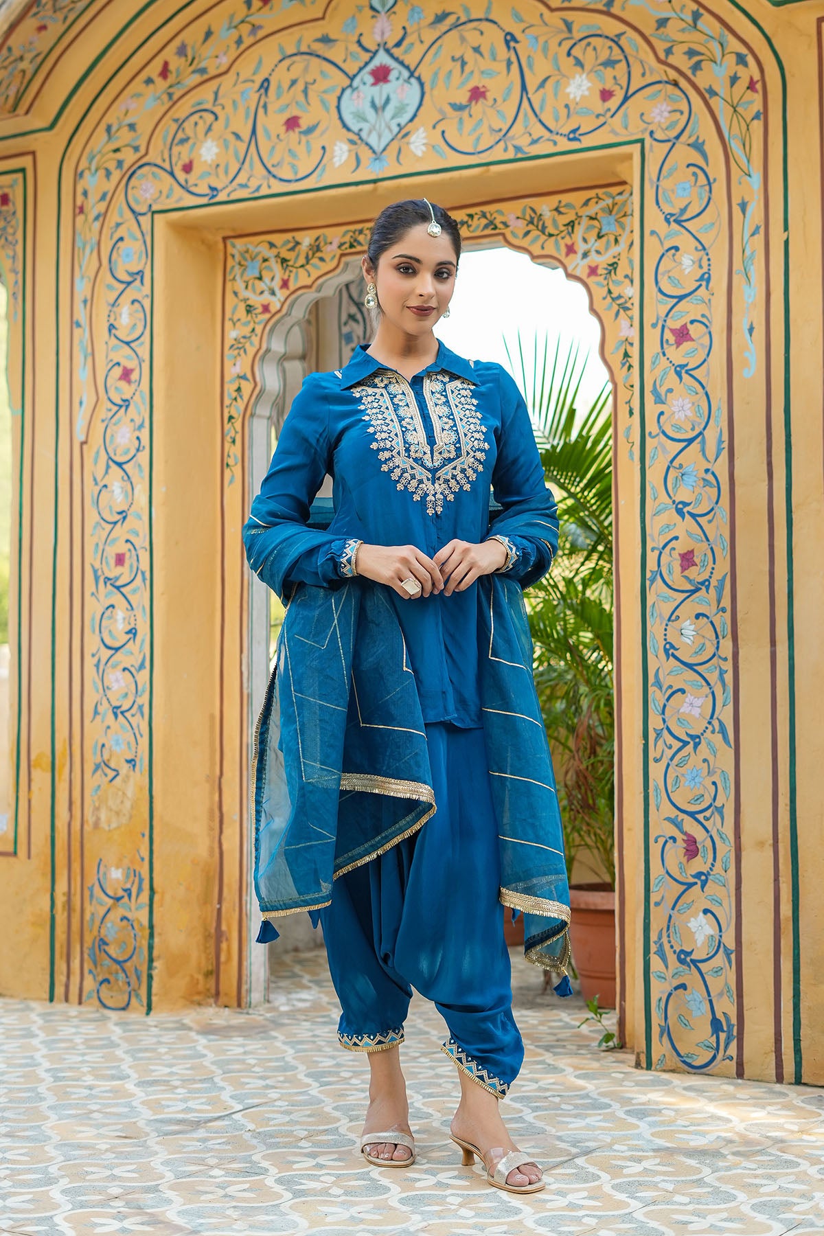 Woman in a blue traditional outfit standing in front of an ornate archway.