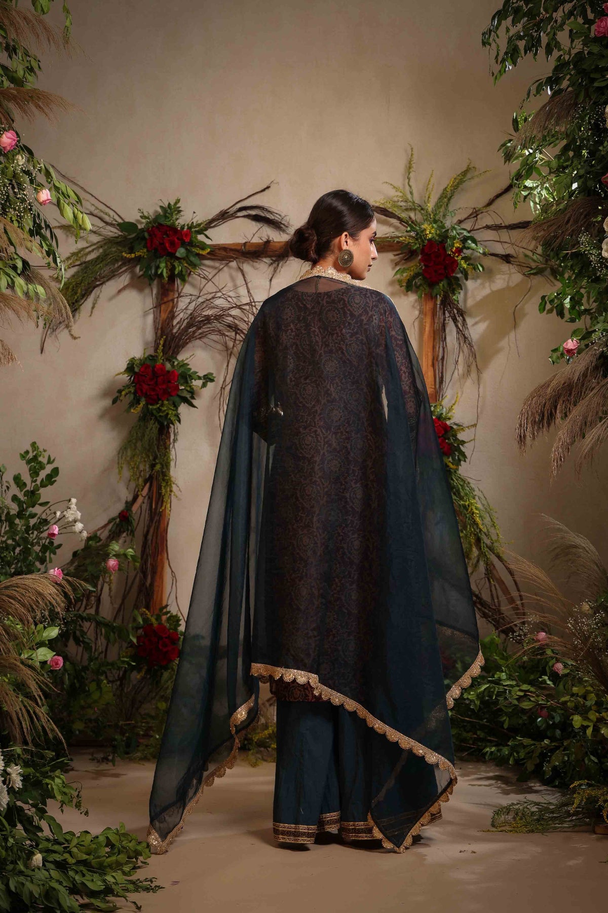 Woman in a dark blue traditional outfit with a cape, standing in front of floral and leaf decorations.
