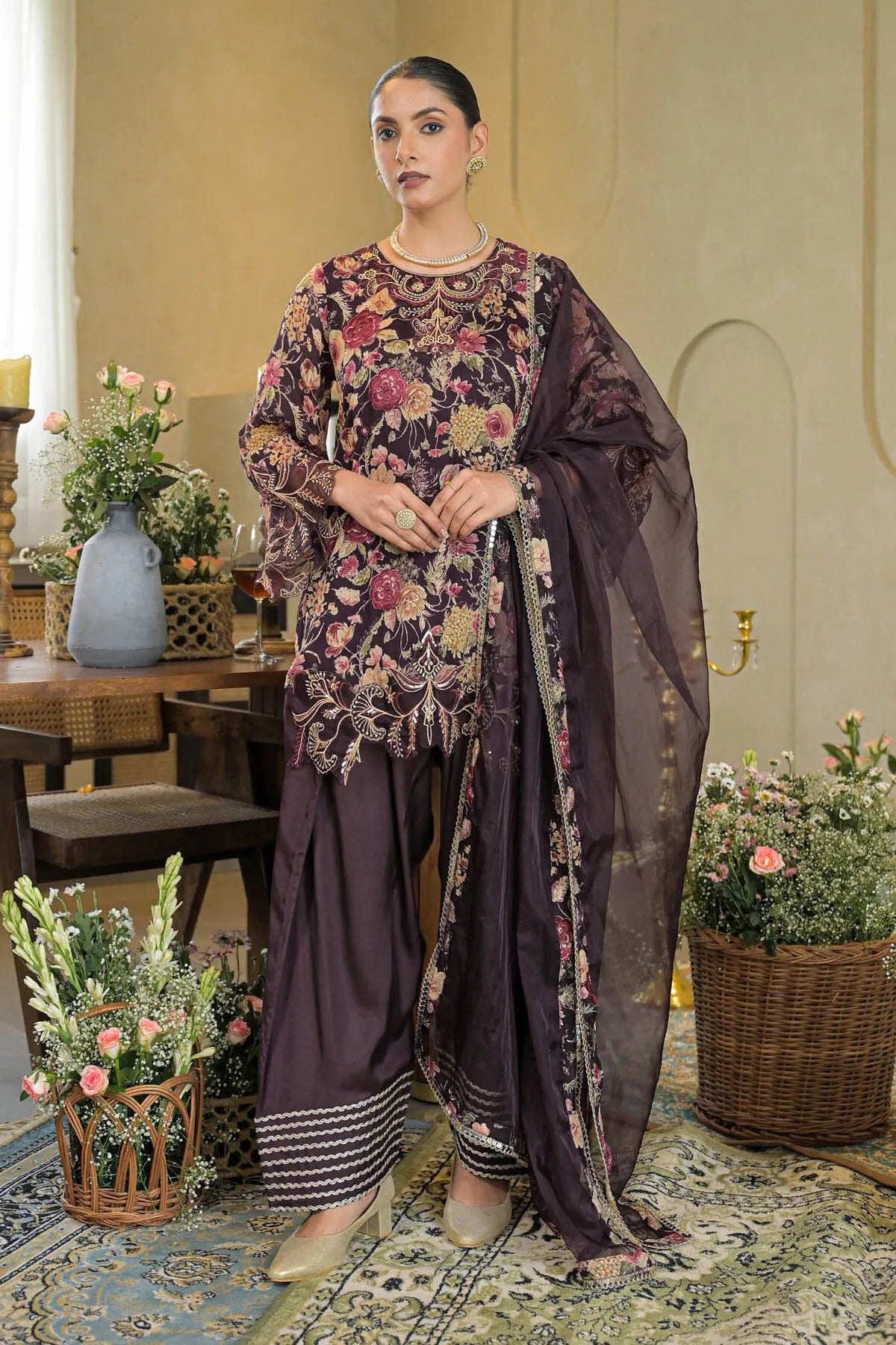 Woman in a floral dress and dupatta standing in an elegant room with decorative plants and furniture.