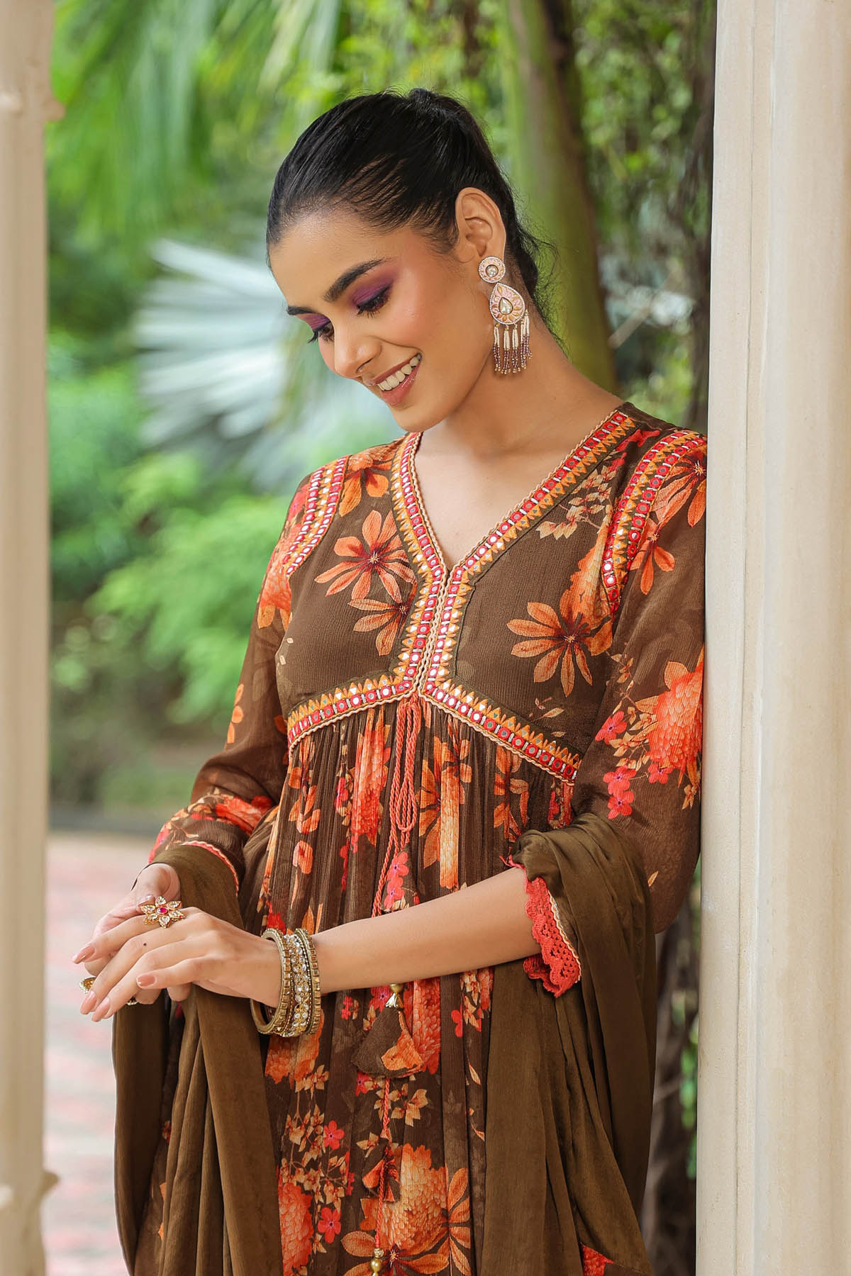 Woman wearing a brown floral dress with intricate patterns, standing outdoors with greenery in the background.