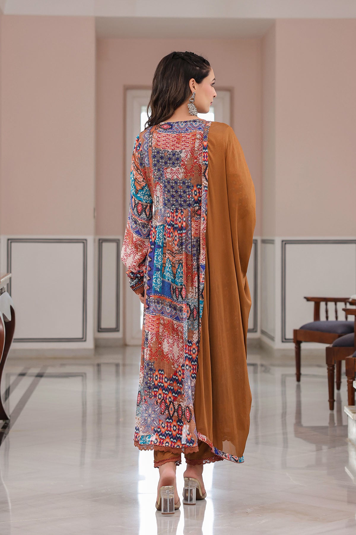 Woman wearing a colorful patterned shirt and brown pants in a room with white walls and wooden furniture.