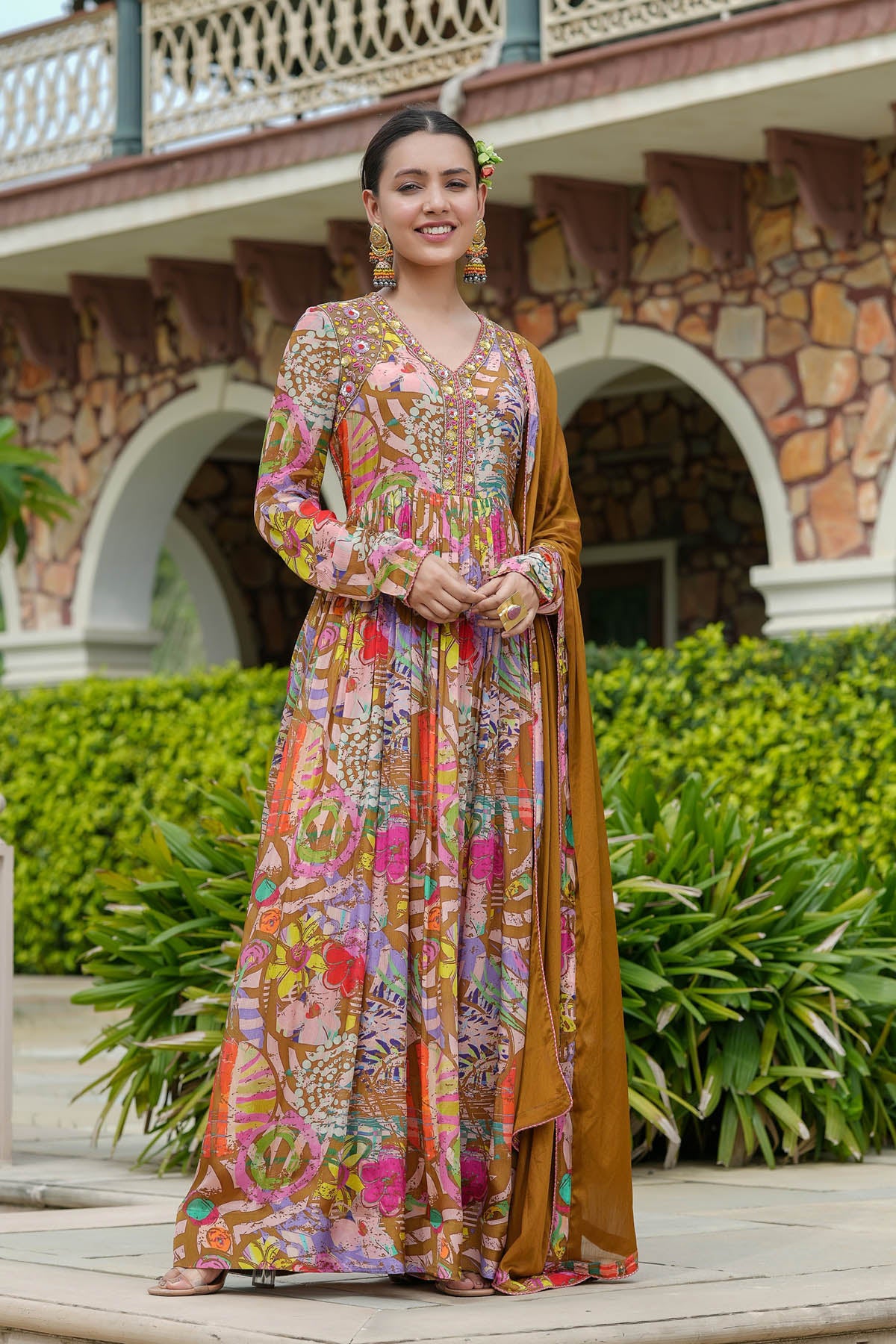 Woman in a colorful traditional outfit standing in front of a stone building with greenery.