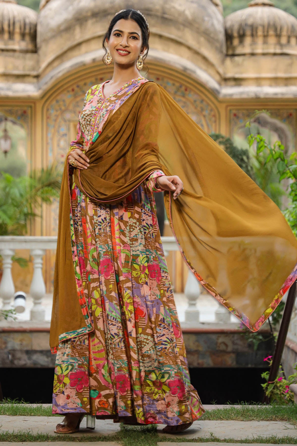 Woman in a traditional outfit with a colorful saree and mustard dupatta, standing in front of an ornate building.