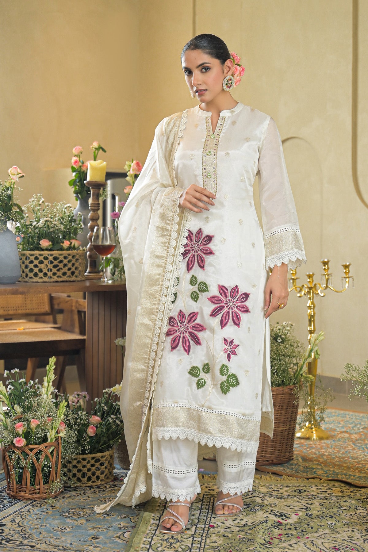 Woman in a white embroidered outfit standing in an elegant indoor setting with floral decorations.