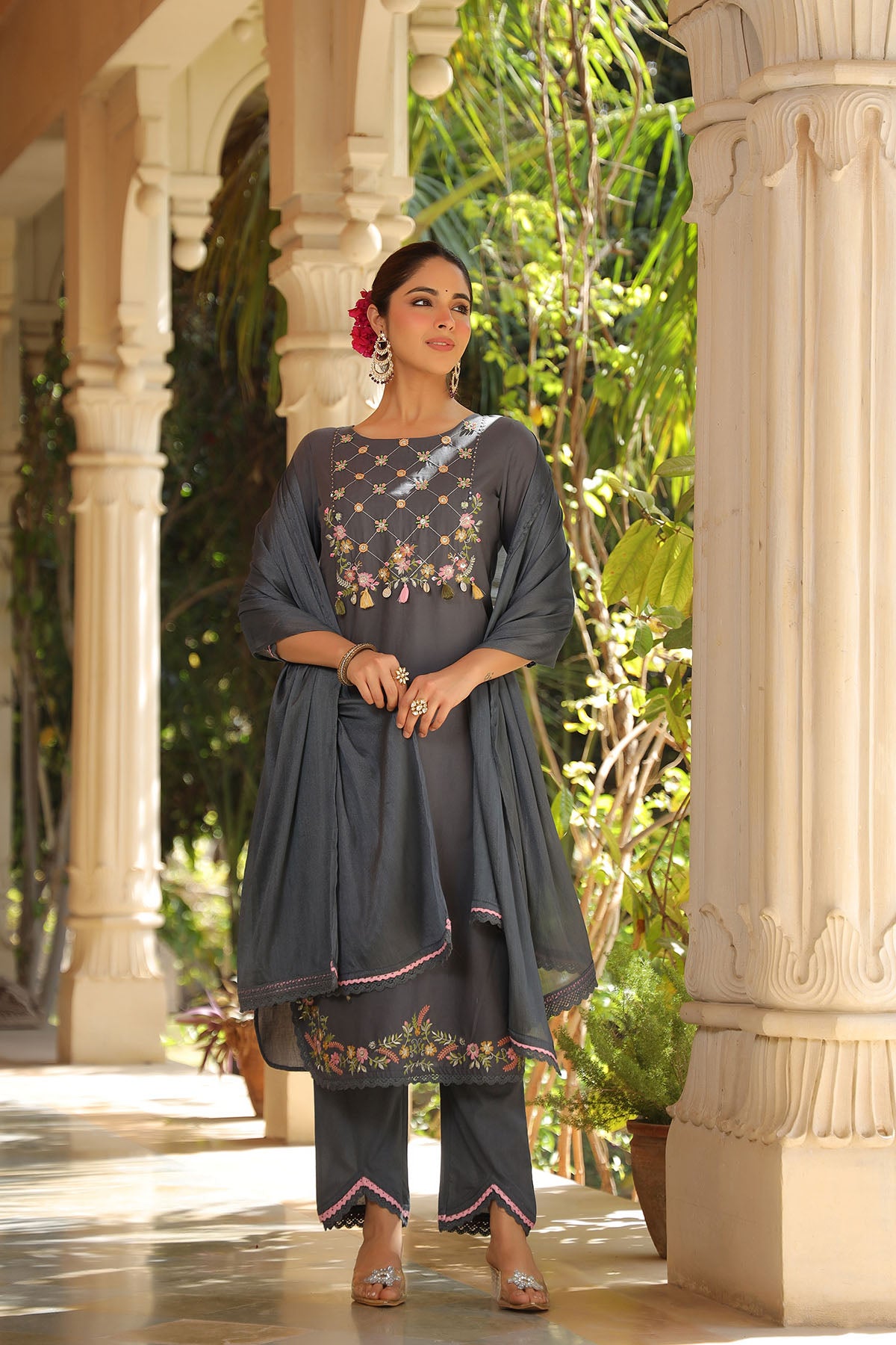 Woman in a traditional outfit standing in an outdoor setting with columns and greenery.