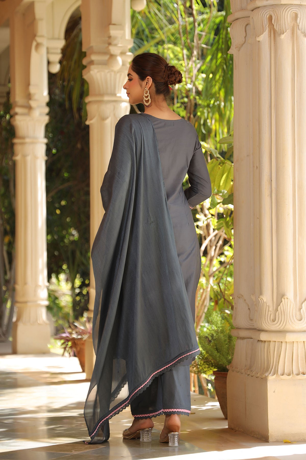 Woman in a gray traditional outfit standing in an outdoor setting with columns and greenery.