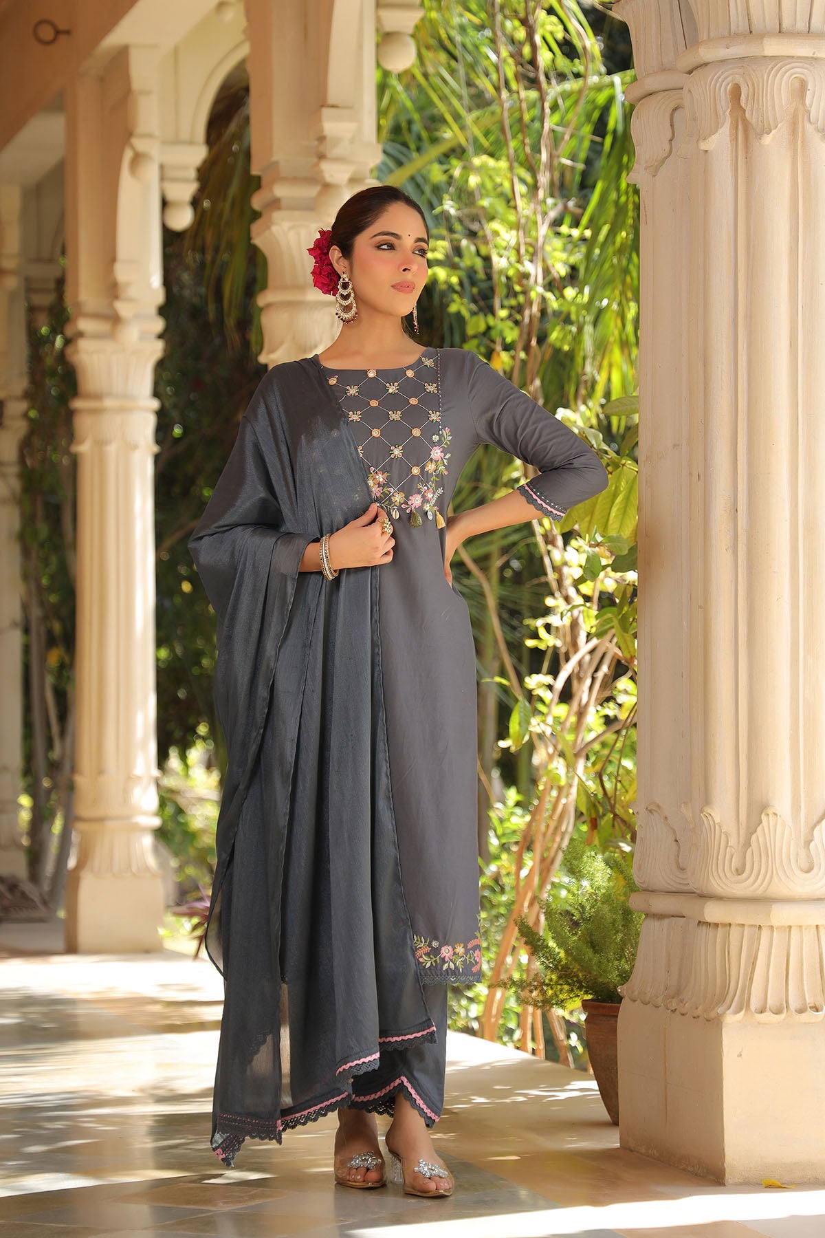 Woman in a gray traditional outfit standing in an outdoor setting with columns and greenery.