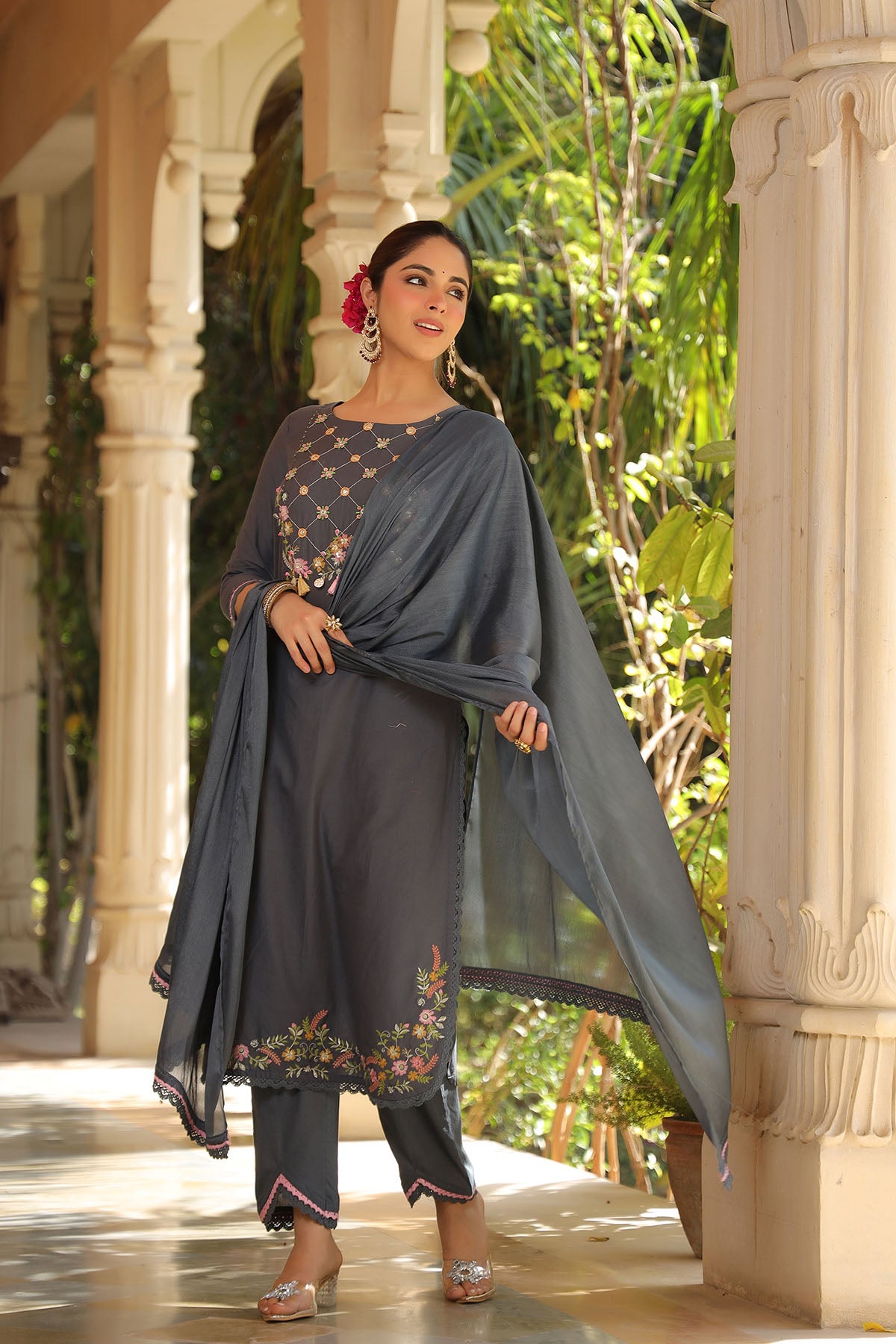 Woman in traditional outfit with floral design standing outdoors near architectural columns.