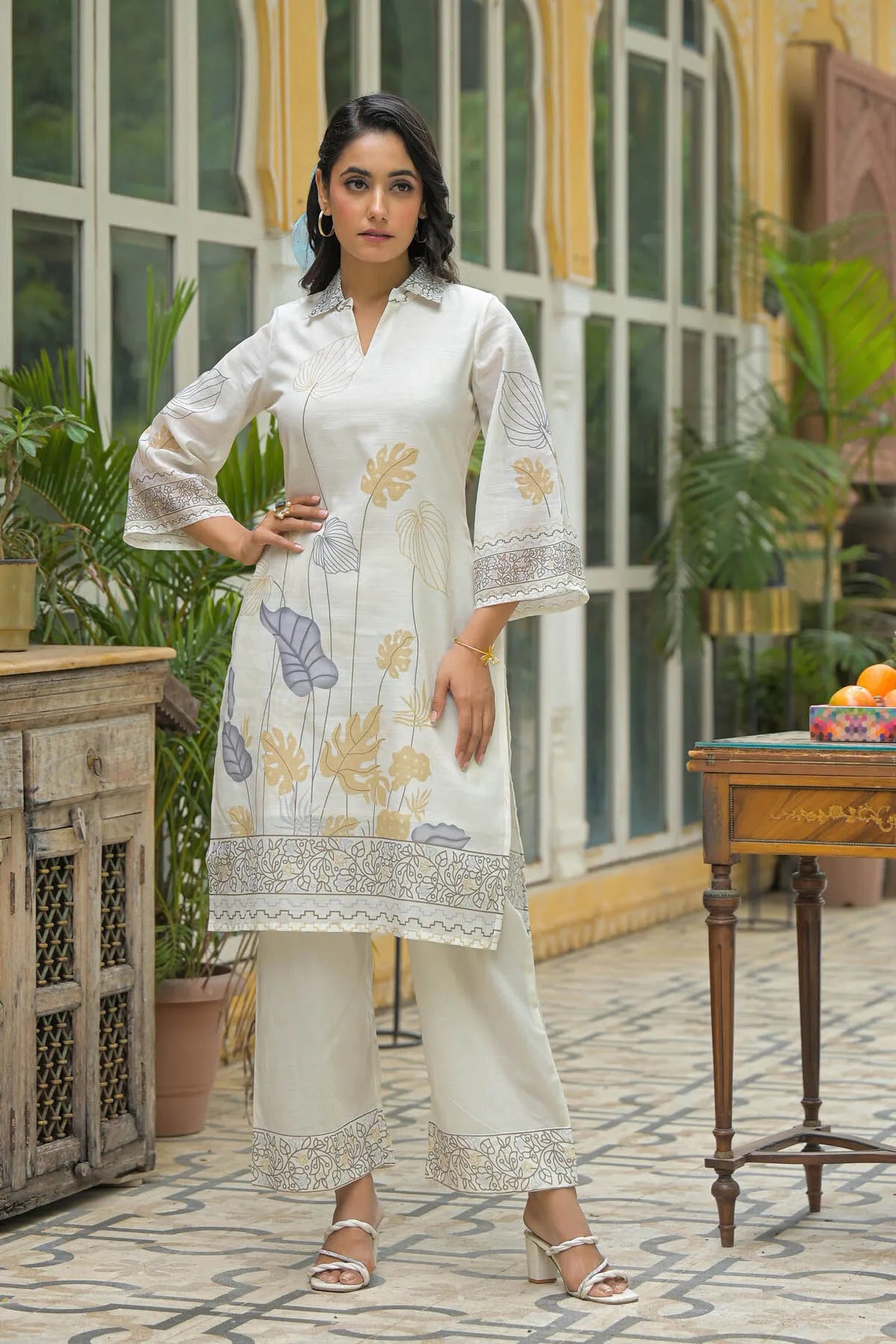 Woman in a white embroidered outfit standing in an outdoor setting with plants and a table.