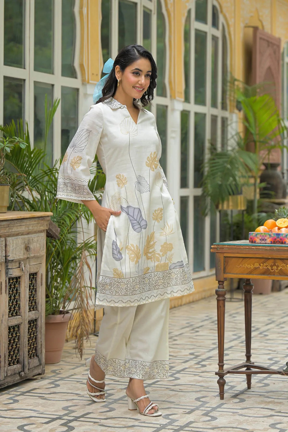 Woman in a white floral dress standing in an outdoor setting with plants and a table.
