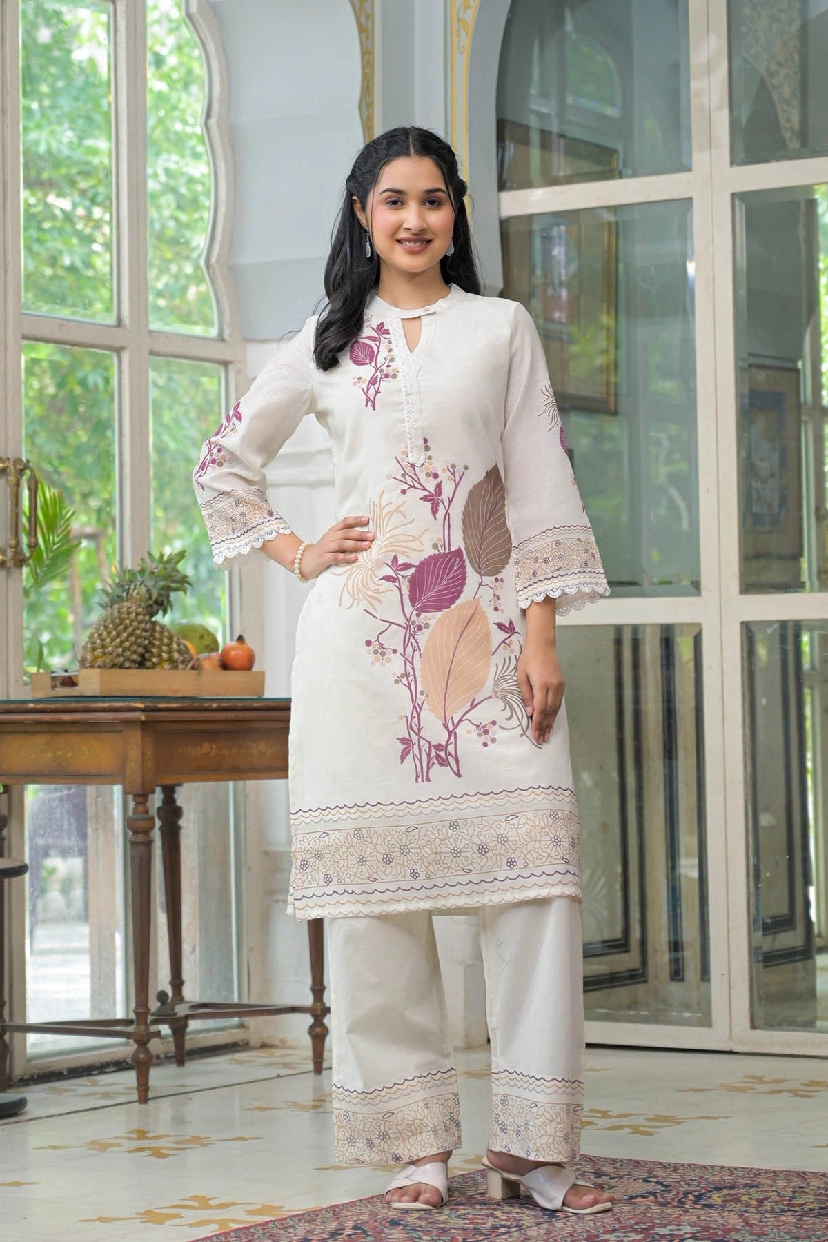 Woman wearing a white embroidered outfit standing in a room with large windows.