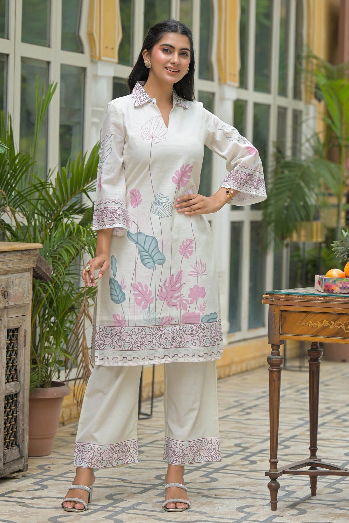 Woman wearing a white floral dress with a patterned border in an outdoor setting.