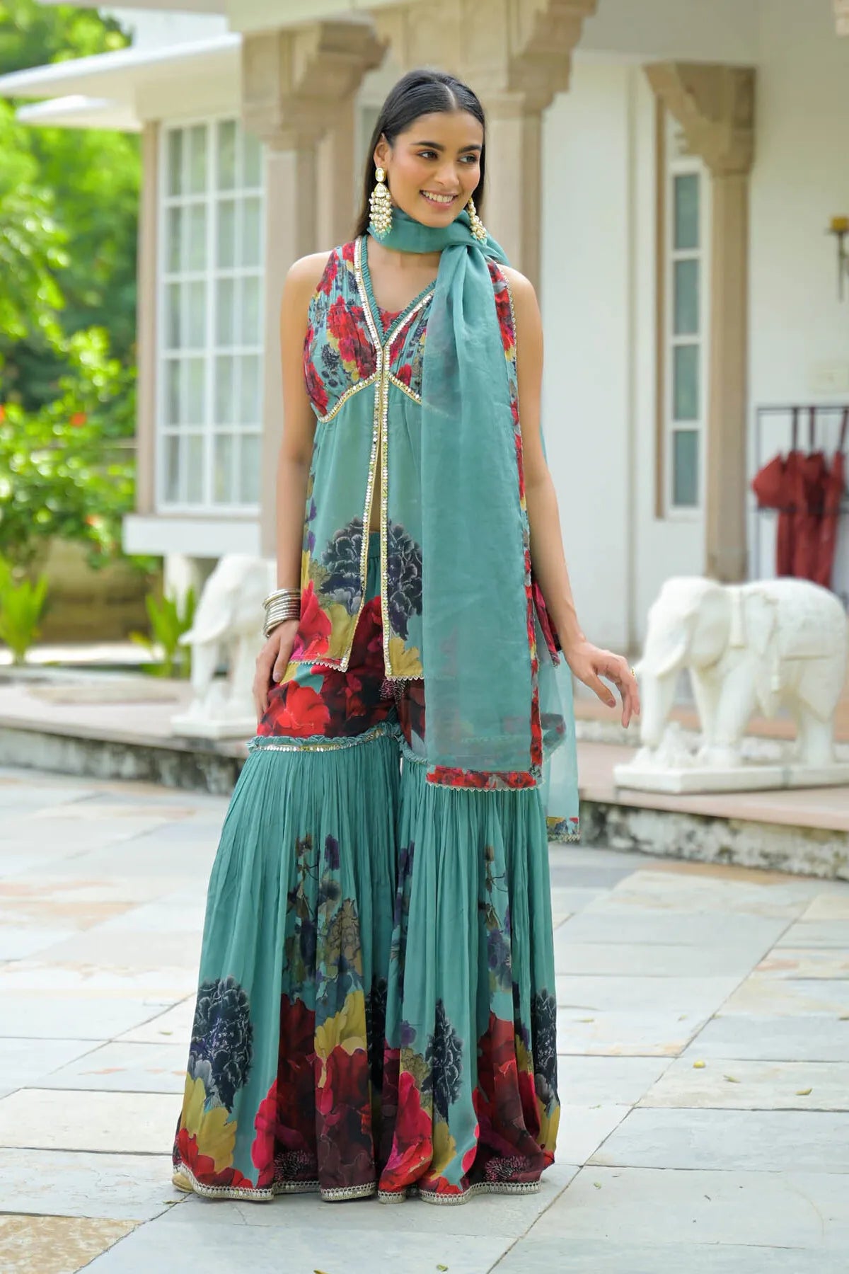 Woman in a colorful traditional outfit standing outdoors with a house and elephant statue in the background.