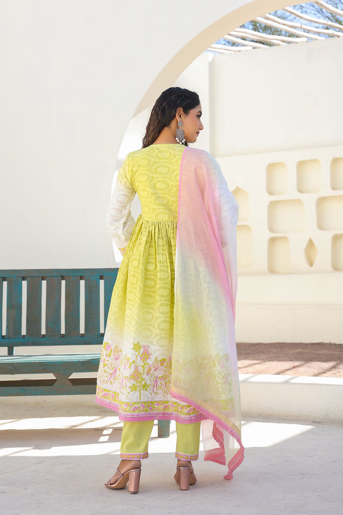 Woman in a yellow traditional outfit with a pink and white dupatta standing in a sunlit room.