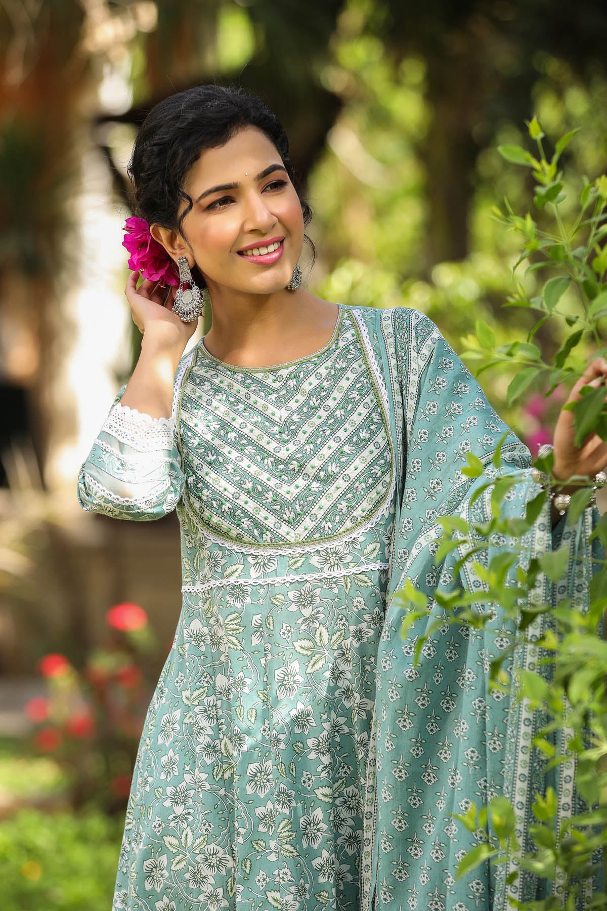 Woman in a green traditional outfit standing outdoors with greenery and flowers in the background
